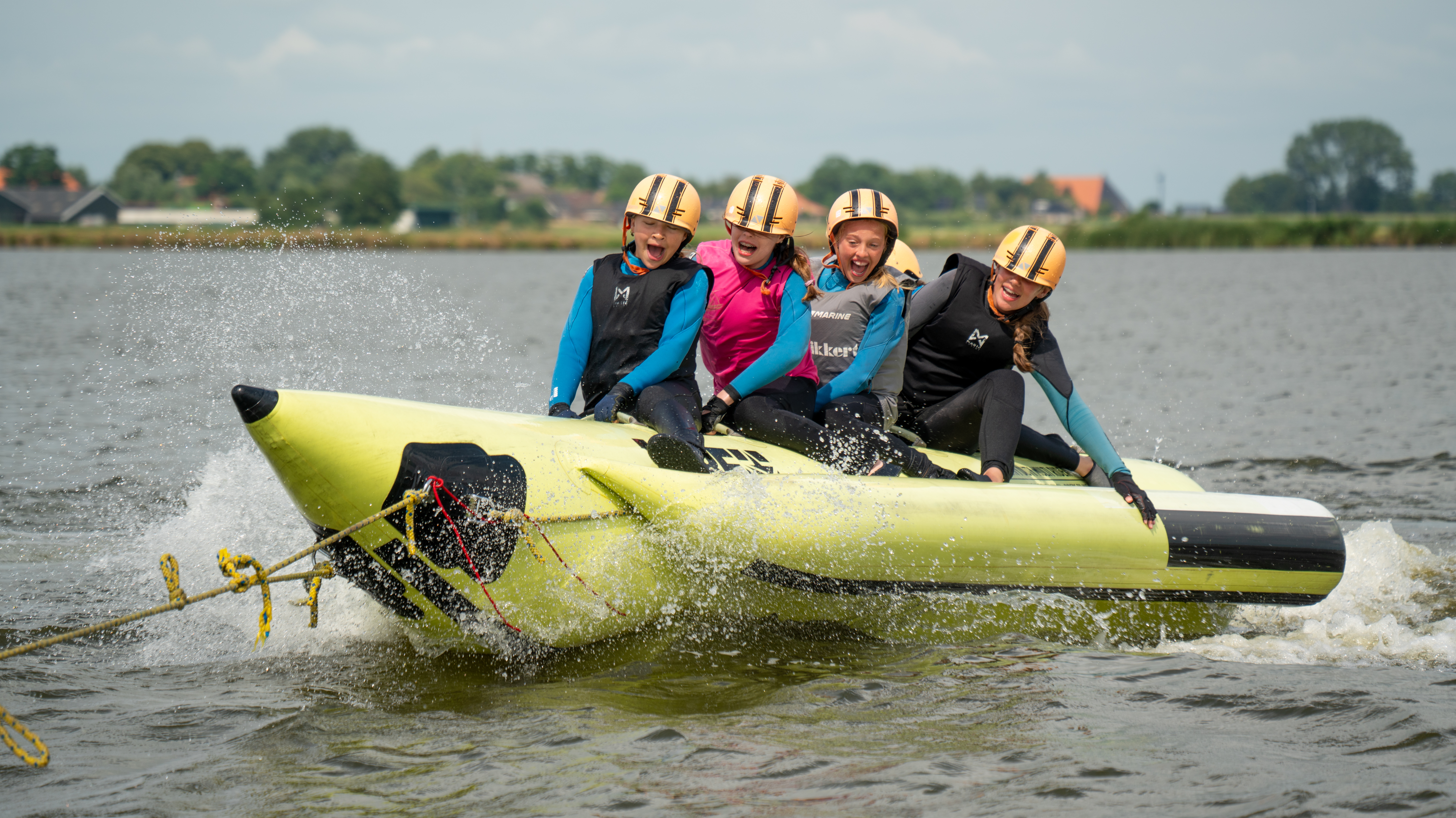 Vier kinderen met helmen en zwemvesten op een gele banaan die door het water raast met een grote spat