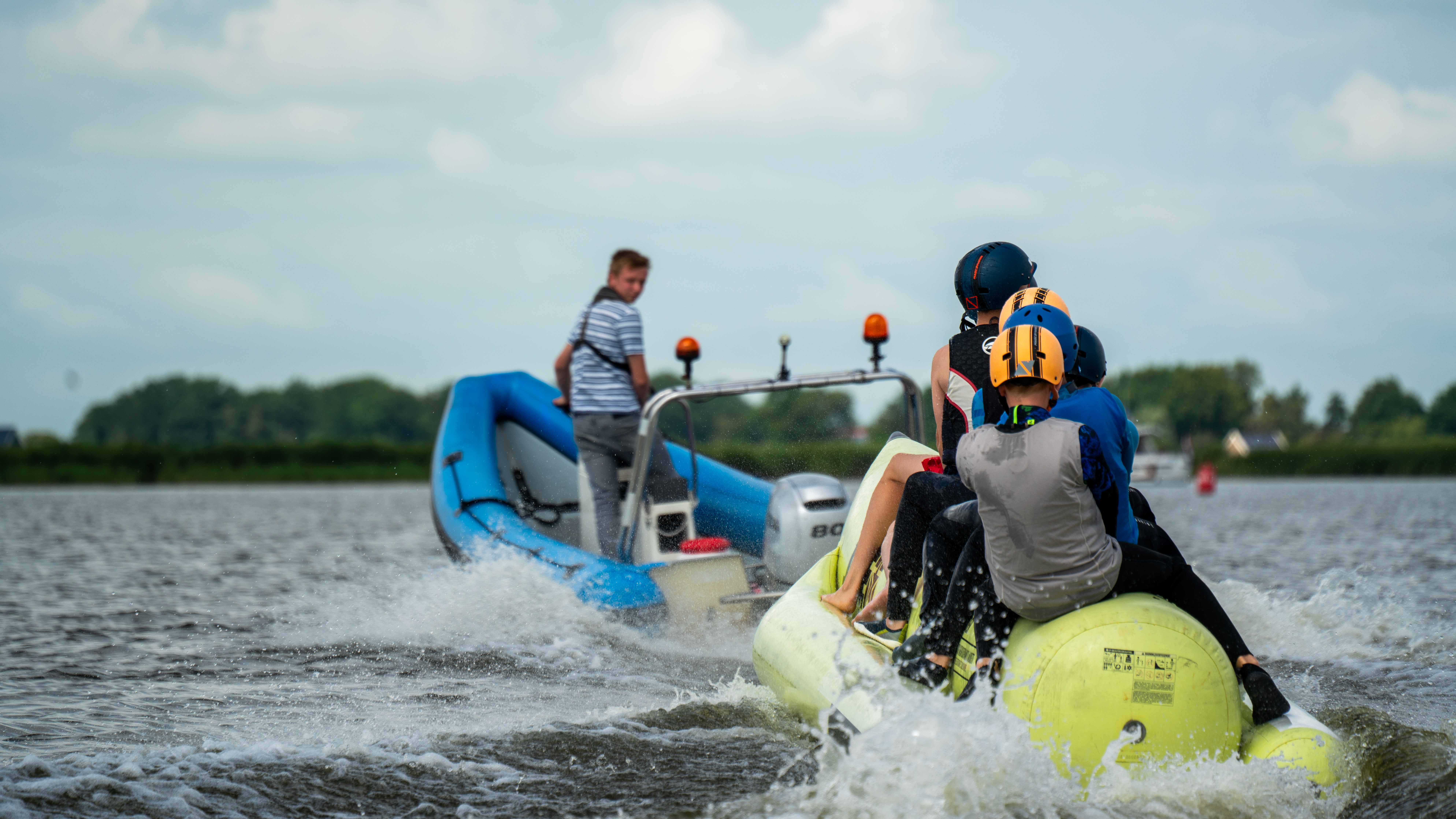 Begeleider in een blauwe motorboot trekt kinderen met helmen en zwemvesten op een gele banaan over het water