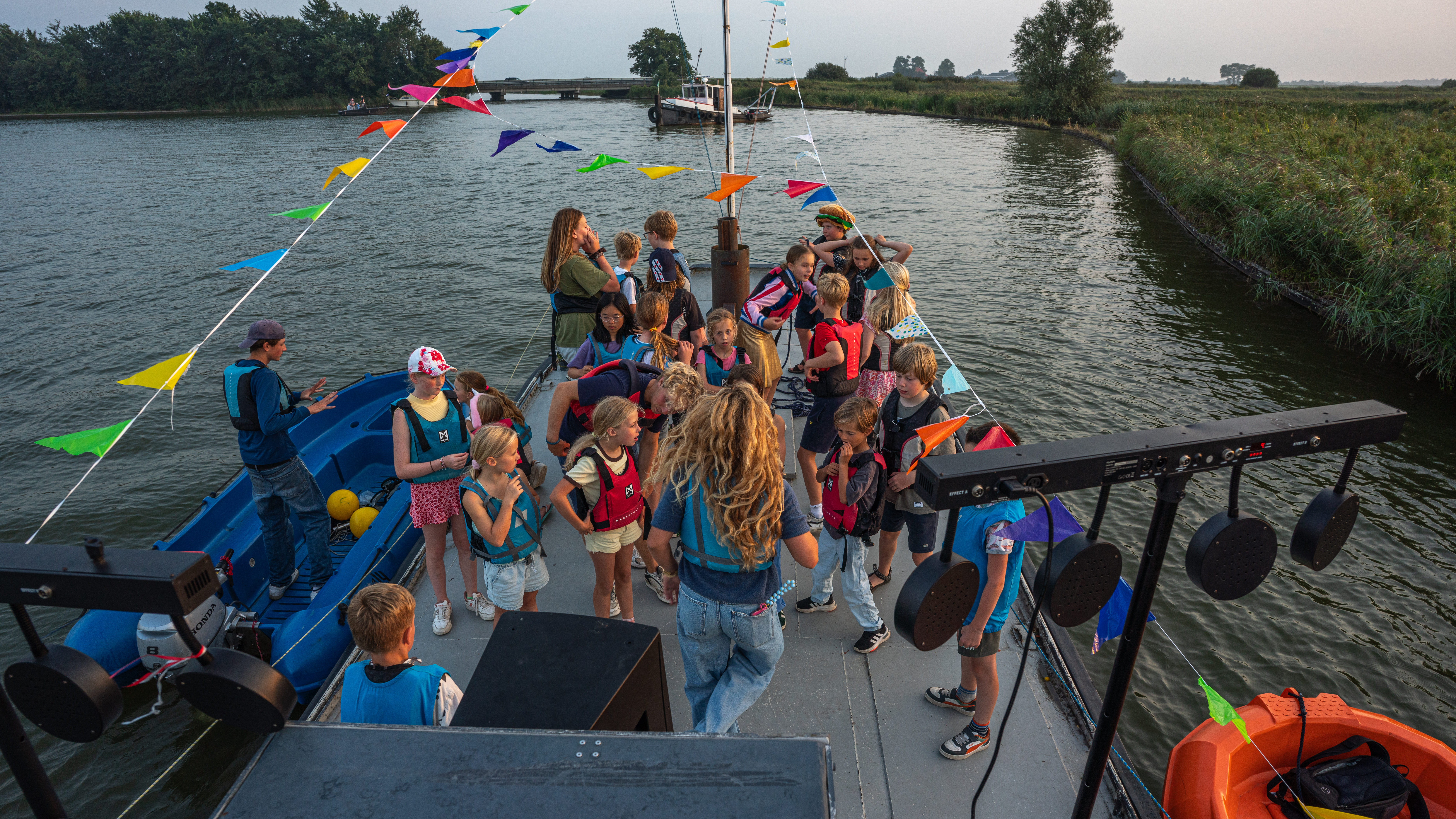 Grote groep kinderen met zwemvesten op het dek van een versierde praam tijdens de schemering