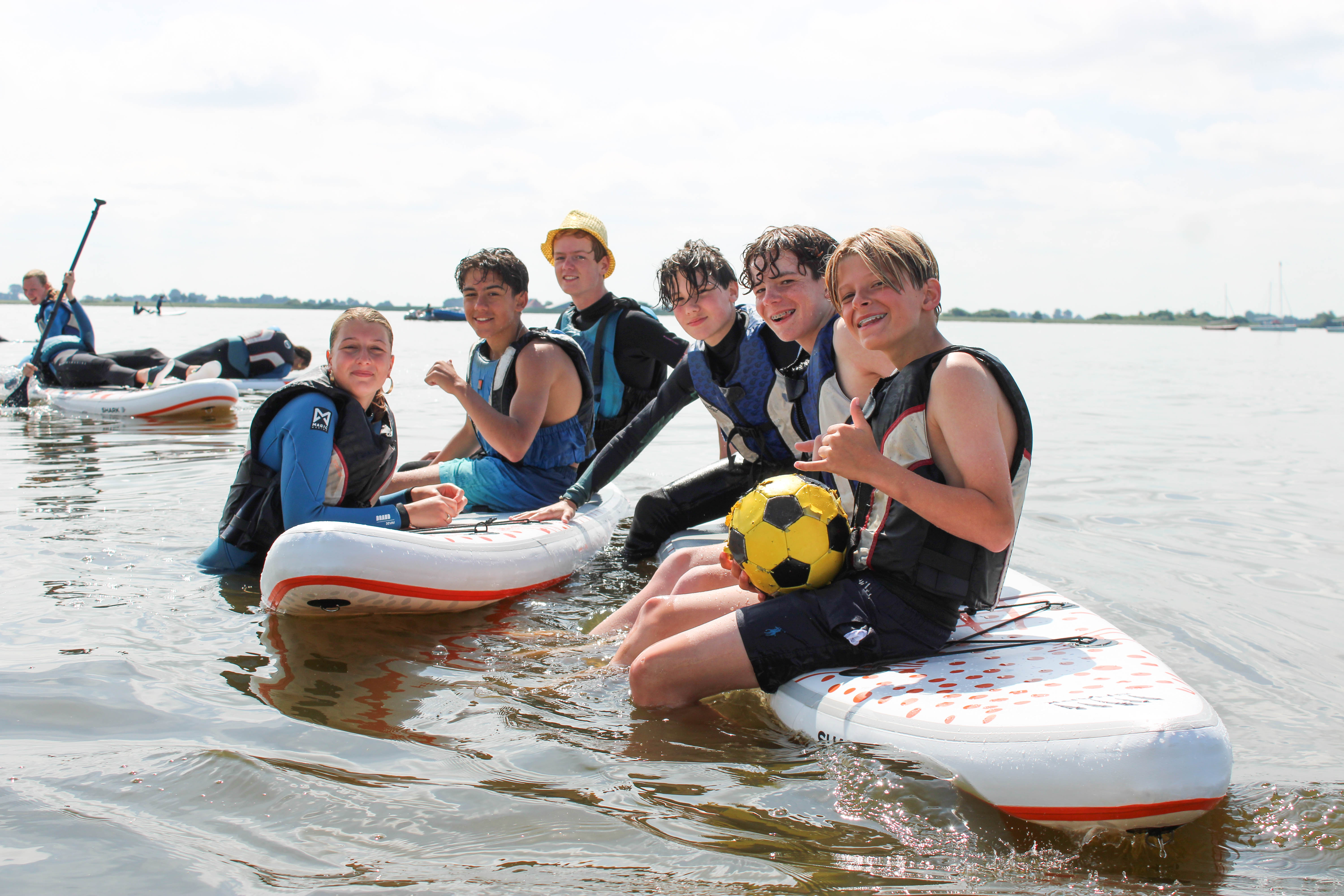 Een vrolijke groep tieners in zwemvesten op supboards op het water, waarbij één jongen een gele voetbal vasthoudt